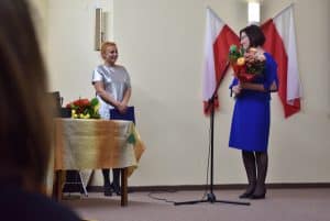 Za mikrofonem stoi kobieta, trzyma w obu rękach bukiet, za nią znajdują się dwie biało – czerwone flagi. Po lewej za stołem stoi kobieta trzymając niebieską teczkę, na stole znajduje się drugi bukiet.