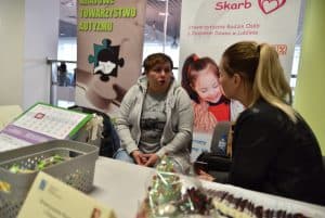 Kobieta siedząca przy stole z rękodziełami. Ich twarze są zwrócone ku sobie nawzajem. W tle banery reklamowe o autyzmie.