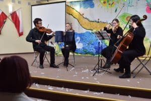 Na scenie znajdują się cztery osoby siedzące na krzesłach, od prawej: mężczyzna i dwie kobiety grające na skrzypcach oraz jedna grająca na wiolonczeli. Przed nimi znajdują się trzy pulpity z kartkami. W tle znajduje się ściana z rysunkiem przedstawiającym: chmury, słońce, jeziorem i łabędziem.