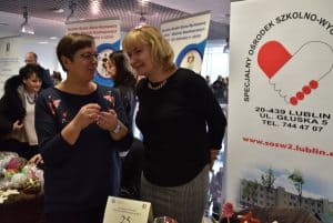 Kobieta nachyla się w stronę drugiej kobiety. Uśmiechają się do siebie nawzajem. W tle banery reklamowe.