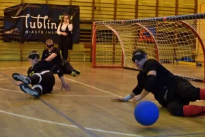 Akcja ma miejsce na sali gimanstycznej. Troje mężczyzn z opaskami na oczach leży na parkiecie. Przy jednym z nich jest duża niebieska piłka, a w tle widać ogromną bramkę i stojącą Panią przy bramce.