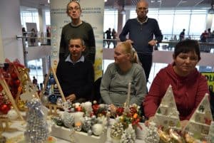 Ludzie siedzący przy stole z rękodziełami bożonarodzeniowymi. w tle dwóch stojących mężczyzn, jeden z nich opiera się o balustradę.