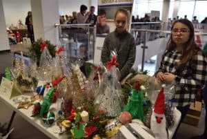 Dwie młode dziewczyny stojące przy stole z rękodziełami. Niektóre wyroby manualne są zapakowane w przezroczysty celofan owinięty czerwonymi kokardami.