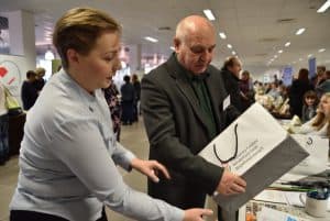 Mężzczyzna trzymający w dłoniach torbę prezentowa. Kobieta obok niego wyciąga przed siebie ręce. W tle inni ludzie siedzący przy stołach.