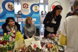 Kobiety siedzą przy stoisku z rękodziełami. Jedna z kobiet stoi i nachyla się w stronę stołu.