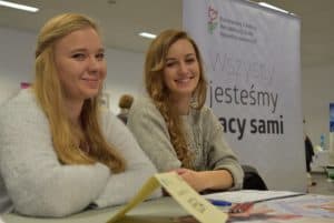 Dwie młode kobiety siedzą przy stole z rękoma założonymi na siebie. Uśmiechają się do zdjęcia.