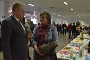 Kobieta stoi i twarz ma skierowaną w stronę stojącego obok niej mężczyzny. Kobieta w rękach trzyma kurtkę. W tle stoły zastawione różnymi robótkami ręcznymi i wokół nich spacerujący ludzie.