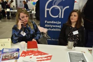 dwie kobiety siedzące przy stole. Jedna z kobiet w dłoniach trzyma telefon, który przykłada do swojej twarzy. Na stole są broszury i ulotki.