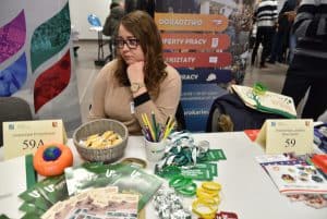 Młoda kobieta siedzi przy stole. Podpiera brodę dłonią. Na stole leżą ulotki reklamowe, długopisy, opaski silikonowe na nadgarstki.