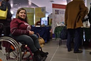 Kobieta na wózku inwalidzkim uśmiecha się do zdjęcia. W tle mężczyzna odwrócony tyłem do zdjęcia. W tle także banery reklamowe pionowe stojące.