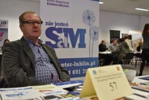 Starszy mężczyzna w okularach siedzi przy stole. Na stole leżą różne ulotki reklamowe. W tle pionowy baner stojący na którym pisze “nie jesteś sam - sm”.