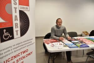 Mężczyzna w wieku średnim siedzi przy stole z ulotkami i broszurami. Ręce ma oparte o stół. W dłoniach trzyma okulary.