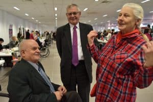 Starsza kobieta uśmiecha się i podnosi ręce ku górze. Obok niej dwóch mężczyzn w garniturach. Jeden z nich stoi drugi siedzi. Uśmiechają się do siebie. Mężczyzna, który stoi ma ręce założone za swoimi plecami. Mężczyzna który siedzi ma dłonie założone na siebie.