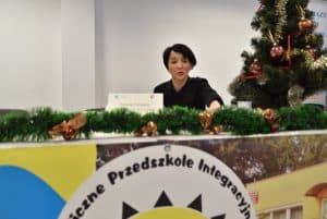 Kobieta siedzi za ladą. Uśmiecha się. Na stole girlanda iglasta. W tle przyozdobiona świątecznie choinka.