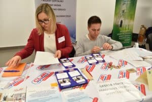 Kobieta i mężczyzna siedzą przy stole. Jedna trzyma w dłoni kartkę. Na stole broszury i ulotki. W tle pionowe banery stojące.