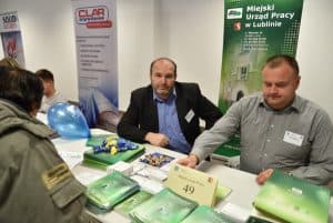 Mężczyźni w garniturach siedzący przy stole z broszurami i ulotkami. W tle banery reklamowe.