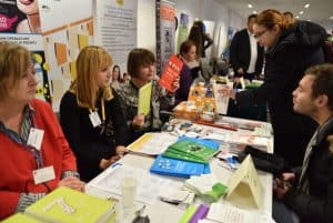 Ludzie siedzą przy stole na którym leżą różne kolorowe broszury i ulotki. Niektórzy nachylają się do tego stołu. W tle pionowe banery stojące.