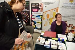 Kobieta siedzi przy stole z broszurami i ulotkami. Jej twarz i wzrok odchyla się w prawą stronę. Po lewej stronie jest kobieta, która nachyla sie do stołu z broszurami. W dłoniach trzyma teczkę.