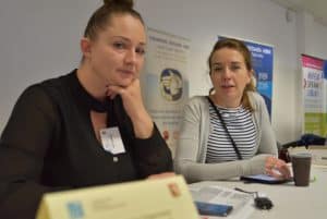 Dwie kobiety siedzące przy stole. Jedna z nich podpiera dłonią swój podbródek. Druga ma położoną dłoń na stole. W tle kubeczek jednorazowy stojący na stole.