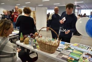 Kobieta siedzi przy stole z rękodziełami i kolorowymi broszurami. Na stole również duży wiklinowy kosz z wyrobami manualnymi. Mężczyzna pochyla się do tego stołu. W dłoni trzyma kartkę. W tle inni ludzie stojący przy innych stoiskach.