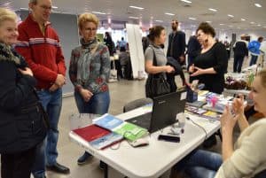 Ludzie stojący przy stole z broszurami. Mają ręce założone na siebie. Na stole stoi również otwarty laptop. W tle ludzie stojący w grupach.