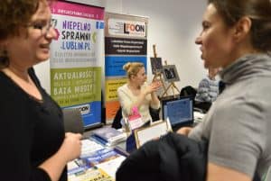 Kobieta siedzi przy stoisku z broszurami. Przy stoisku stoją kobiety twarzami zwróconymi do siebie. Jedna z nich uśmiecha się do drugiej.