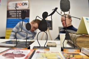 Na stole stoją statuetki ludzików. Mają ręce wzniesione ku górze. W tle mężczyźni siedzący przy stole. W oddali baner reklamowy.