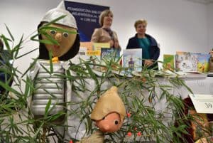 Są figurki ludzików. Jeden z nich ma ubraną na sobie zbroje. Ludziki stoją w liściach rośliny zielonej. W tle dwie kobiety stojące przy stole z rozłożonymi różnymi kolorowymi ksiażkami.