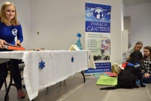 Kobieta stojąca przy stole nakrytym białym obrusem. Kobieta ta nachyla się i trzyma w dłoniach teczkę. Po prawej stronie dwie kobiety siedzą na podłodze razem z czarnym dużym psem.