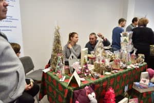 Mężczyzna i kobieta siedzą przy stole z rękodziełami i wyrobami manualnymi, świątecznymi. Na stole choinki, świece, stroiki. W tle ludzie stojący w grupie.