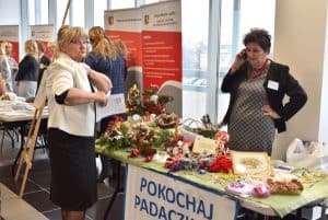 Kobiety stojące przy stole z rękodziełami. Na kancie stołu zawieszony jest baner “pokochaj padaczkę”. Jedna z kobiet trzyma w dłoni telefon przyłożony blisko do twarzy. Druga ręka jest założona na biodra.