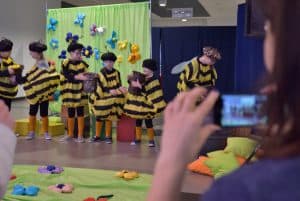 Na scenie stoją dzieci przebrane za pszczoły. W tle kobieta trzymająca telefon w dłoni na wysokości swojej twarzy.