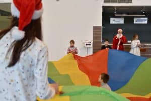 Kobieta z czapką mikołaja trzyma razem z dziećmi kolorową chustę animacyjną. Jedno z dzieci siedzi na środku chusty.