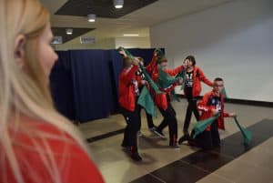 Dzieci stojące na środku sali przybierają różne figury artystyczne. Dzieci ubrane są w czerwone bluzy sportowe, a w dłoniach trzymają zielone szarfy.