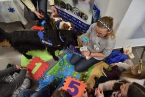 Kobieta siedzi na ziemi. Wokół niej siedzą dzieci i stoją psy. Kobieta trzyma w ręce kolorową kostkę.