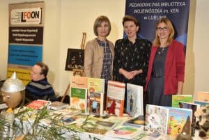 Kobiety stoją przy stole z wystawionymi książkami. Uśmiechają się do zdjęcia. W tle baner reklamowy na którym pisze “Pedagogiczna biblioteka wojewódzka im. KEN w Lublinie”.