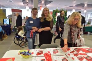 Kobieta obejmuje ramieniem młodego mężczyznę. W tle kobieta siedząca na wózku inwalidzkim. na dole zdjęcia jest stół z cukierkami i kolorowymi broszurami nad którymi nachyla się starsza kobieta.