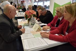 ludzie siedzący przy stole naprzeciwko siebie. Dwie kobiety mają ułożone dłonie na stole. Na przeciwko nich siedzi mężczyzna, który w dłoni trzyma długopis.