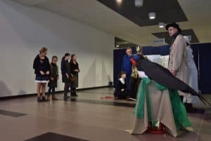 Ludzie stojący na środku sali, przebrani w różne stroje. Jest młody mężczyzna z koroną na głowie. Jest mężczyzna z czarnym kapeluszem i długim brązowym płaszczem. Jest rękodzieło wrony zrobione z bibuły.