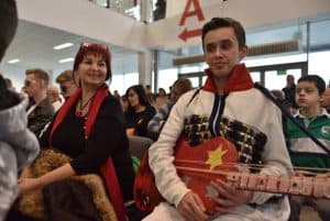 Mężczyzna trzyma w dłoniach kolorową gitarę. Obok niego siedzi kobieta i uśmiecha się do zdjęcia. W tle inni ludzie siedzący na krzesłach.