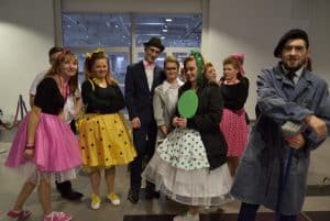 Ludzie stojący w pomieszczeniu ubrani są w kolorowe stroje. Kobiety mają na sobie kolorowe długie spódnice. Mężczyźni są w garniturach kapeluszach i beretach. Wszyscy uśmiechają się do zdjęcia.