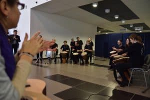 Na środku sali siedzą ludzie na krzesłach i między nogami mają bębenki afrykańskie. Po lewej stronie zdjęcia jest kobieta która ma założone na siebie dłonie.