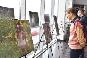 Mężczyzna patrzy na zdjęcia umieszczone na sztalugach. W tle inni ludzie i duże wysokie okna przez które wpada światło do sali.