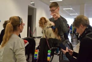 Młody mężczyzna siedzi na wózku inwalidzkim i trzyma na kolanach psa. Wokół niego ludzie twarzami zwróconymi w jego stronę.