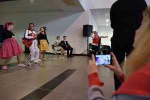 Ludzie w kolorowych strojach stoją na środku sali. Jeden mężczyzna trzyma w dłoniach przewieszona przez swój tułów gitarę. Jedna kobieta wyciąga swoje dłonie ze smartfonem na wysokości swojej twarzy.