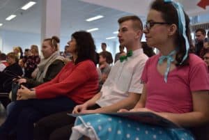 Ludzie siedzący na krzesłach patrzący się przed siebie. Po prawej stronie zdjęcia siedzi kobieta ubrana w różowo niebieski strój. Ma na włosach niebieską kokardę.