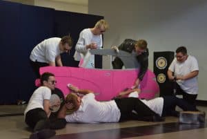 Ludzie są na scenie. Dotykają dużego rysowanego i wyciętego auta. Niektórzy z nich leżą na podłodze i dotykają auta gąbeczkami.