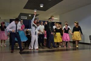 Ludzie stojący na środku sali w kolorowych strojach. Jeden z mężczyzn podnosi jedną rękę ku górze.