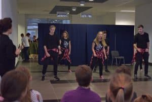 Młodzi ludzie stojący na scenie. Są ubrani w czarne stroje i przewieszone czerwone w kratkę koszule przez biodra. w tle ludzie twarzami zwróconymi w ich kierunku.