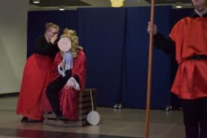 Kobiety są na scenie. Jedna z nich siedzi na drewnianym pudle. Jedna pochyla swoją twarz ku niej i przykrywa usta swoimi dłońmi.